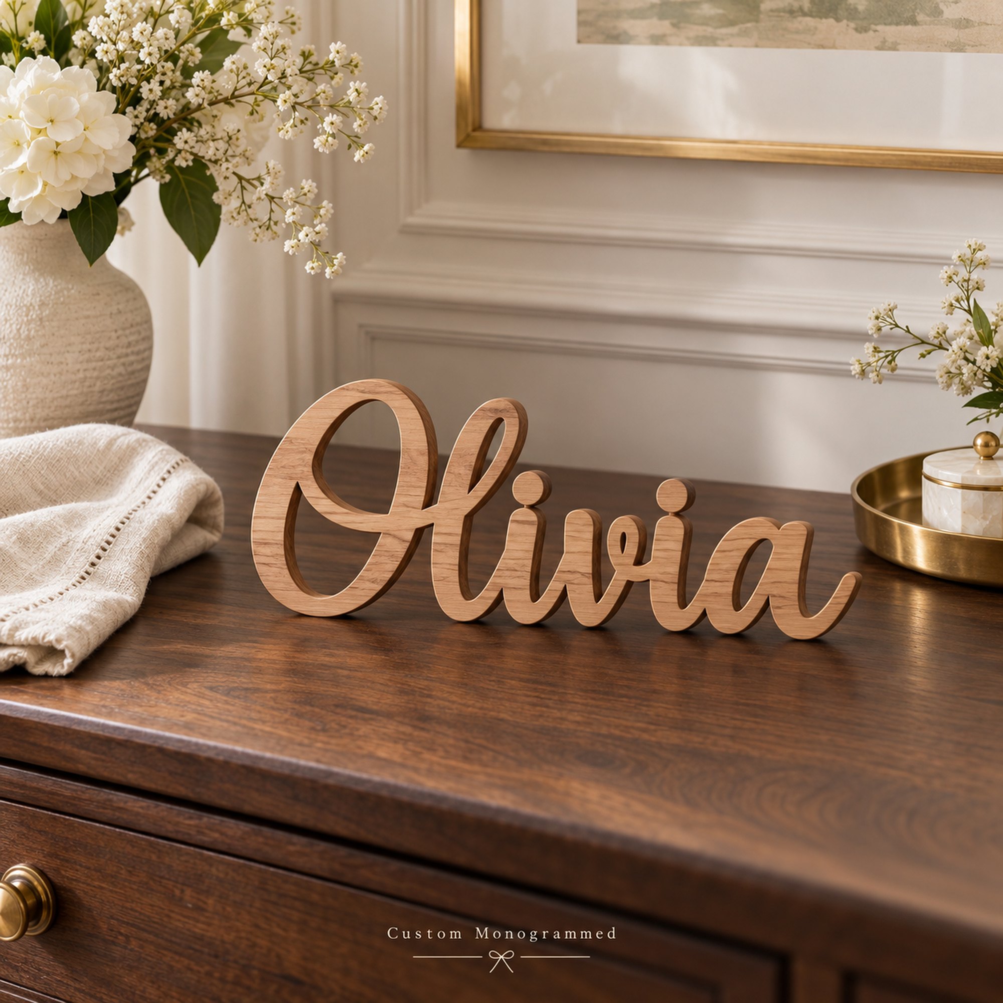 Wooden name sign reading Olivia in script standing on a walnut console beside a vase of white flowers and a brass tray.